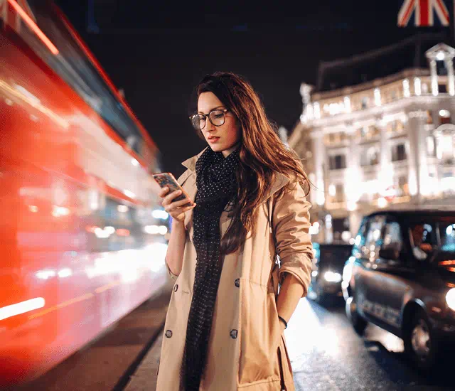woman wearing glasses, black scarf and tan coat looking down at phone next to blurred, busy street